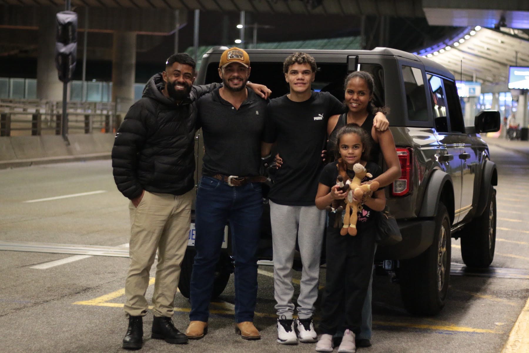 Cinco pessoas, entre elas uma criança, sorrindo em frente a um carro, à noite, em um aeroporto